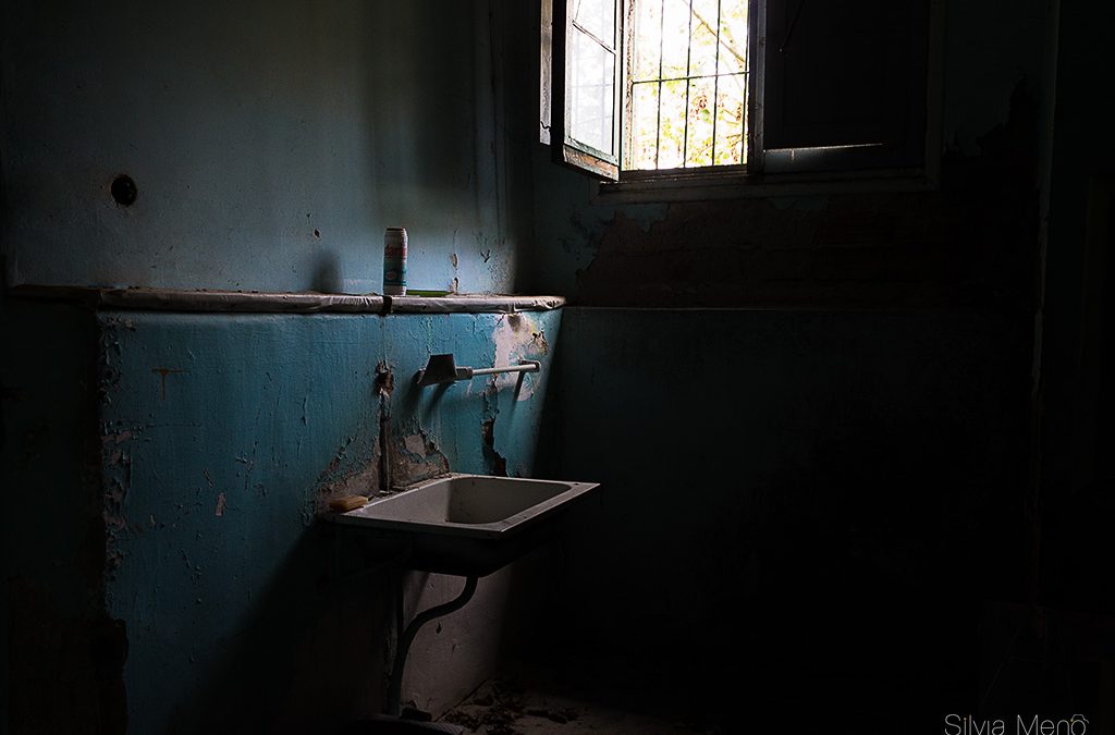 Un baño de una casa abandona, la pared es azul y parte de la pintura se ha caído , se ve un lavabo sin grifo y una ventana de madera vieja por lo que entra un poco de luz.