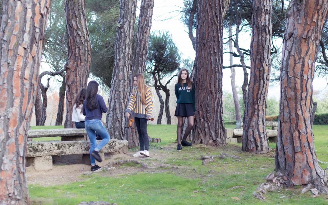 En un parque con muchos árboles se observan tres niñas hablando entre ellas junto a una mesa y otra niña en el fondo mirando a cámara apoyada en un árbol.