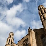 Parte de la fachada de una iglesia con dos torres en los lados y un cielo azul con muchas nubes.