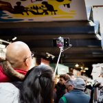 Una pareja se está sacando una foto con un móvil sujetado con un palo selfi. En el fondo se ve mucha gente y una serie de puestos.