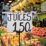 Se ve una mesa grande llena de zumos preparados y un cartel que pone "JUICES 1.50€", al fondo se ve un mostrador lleno de fresas enormes.