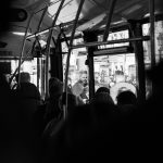 En el interior de un autobús se observa mucha gente en sus asientos, en el fondo se ve gente transitando en la calle.