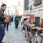 En una calle se ve a un hombre observando bicicletas antiguas en un puesto con muebles en la calle.